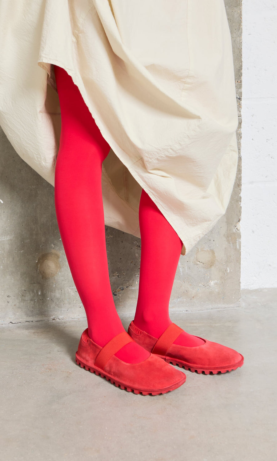 Plümo- Person wearing a cream skirt, Sola Red Flats, and bright red tights, standing on a concrete floor.
