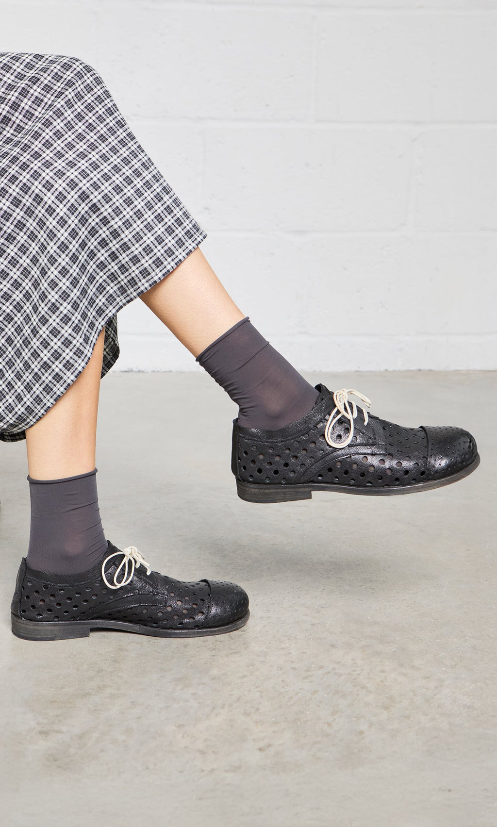 Plümo- Model in Sheer Grey Socks and black perforated shoes with white laces, wearing a plaid skirt, lifts one leg.