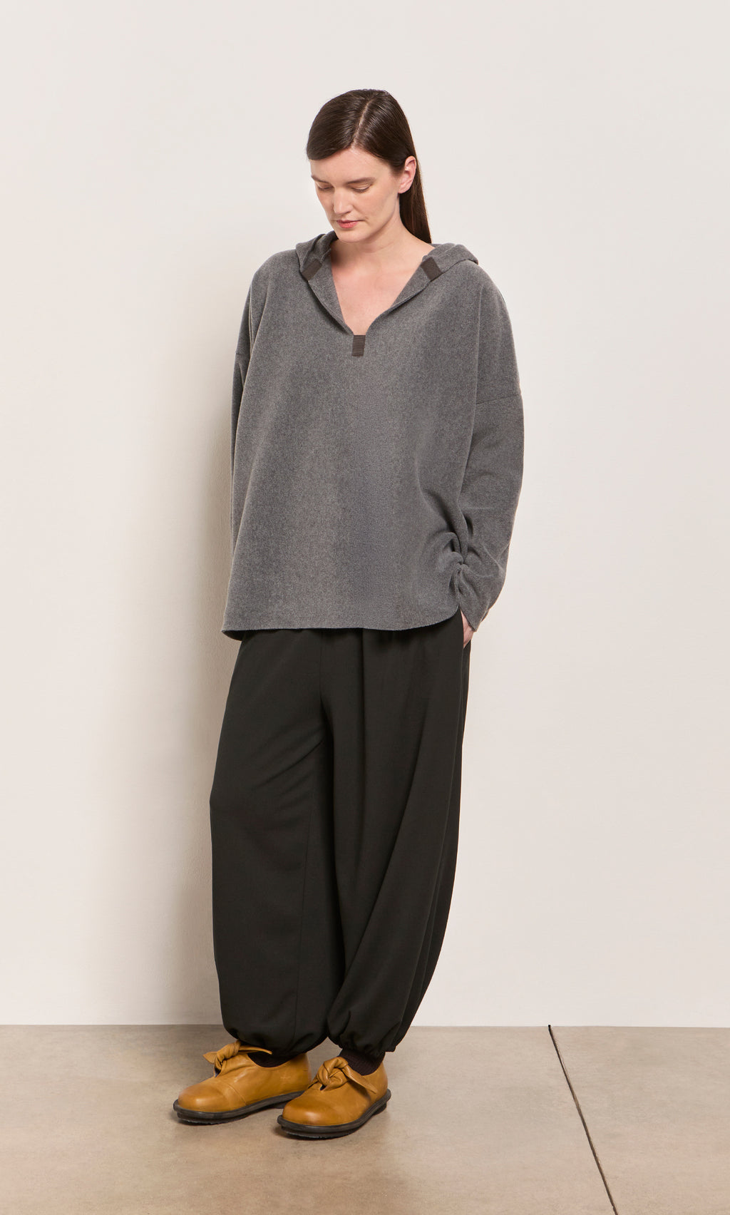 Plümo  Woman in a loose gray hoodie, Mia Balloon Pants, and yellow shoes stands against a plain light background.