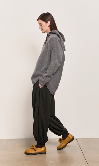 Plümo  Person in a Grey Felted Hoodie and black pants stands sideways, wearing yellow shoes.