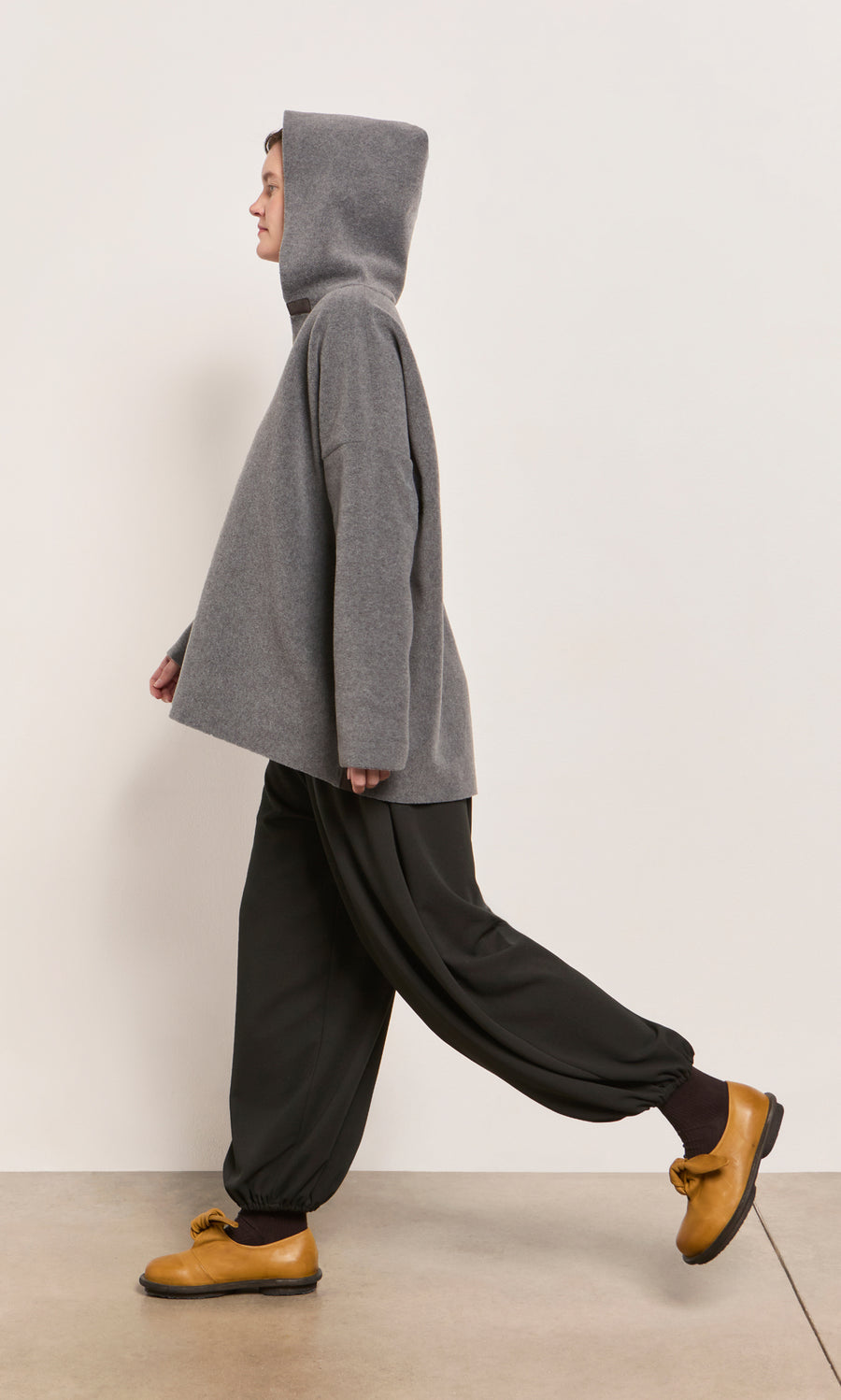 Plümo  Person in a Grey Felted Hoodie, black pants, and yellow shoes walks beside a plain light wall.
