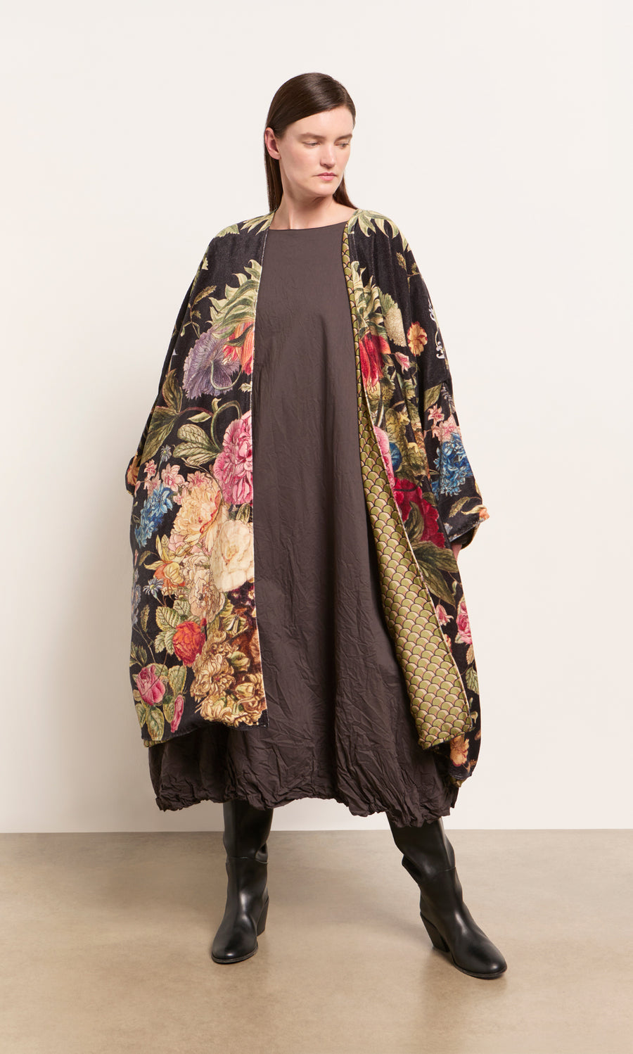 Plümo  Woman in Shilling Velvet Coat, long brown dress, and black boots stands against a plain, light-colored wall.