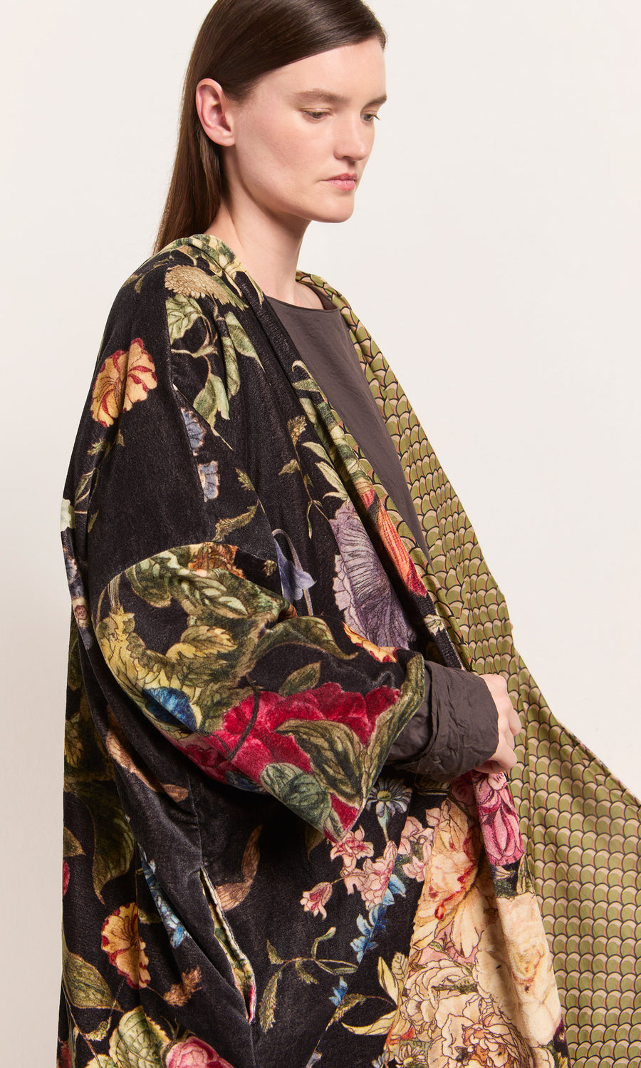 Plümo  Woman in a Shilling Velvet Coat over a brown top, standing against a plain background, looking to the side.