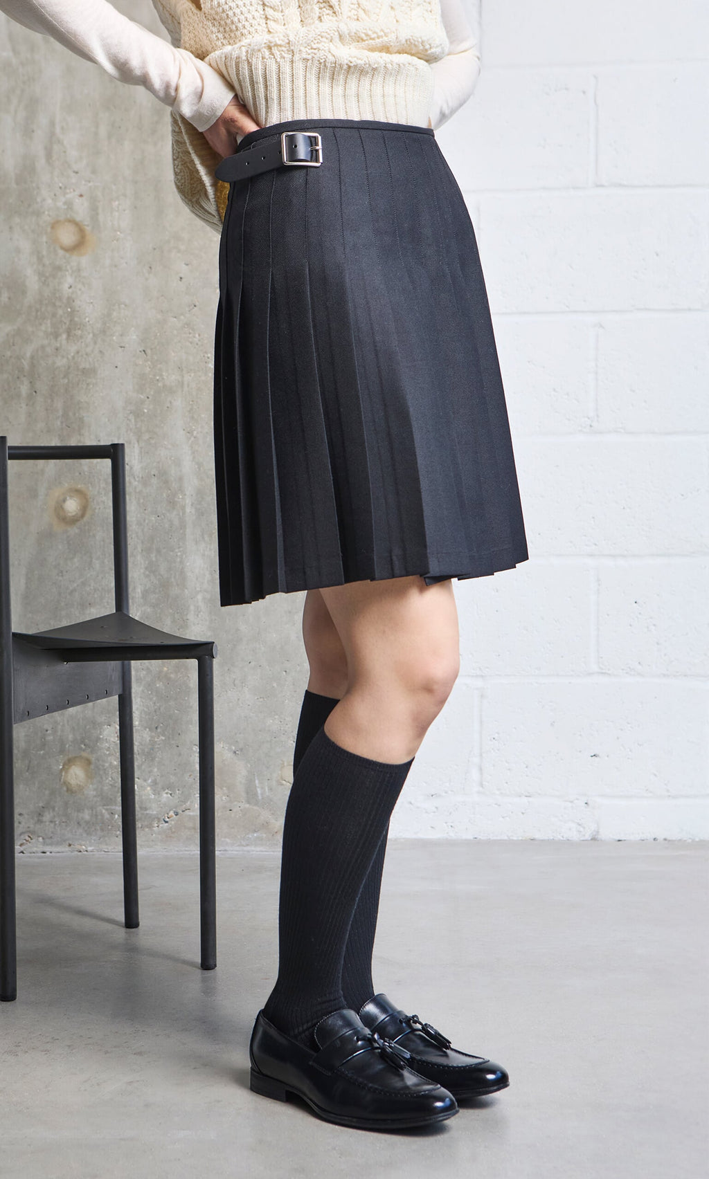 Plümo- Person wearing a Short Black Kilt stands by a black chair in a modern room; kilt made in Ireland.