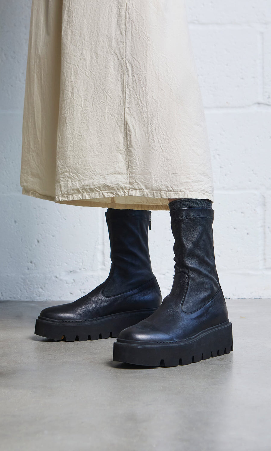 Plümo- Person in a long cream skirt wears Short Prato Boots with chunky soles, standing on a concrete floor.