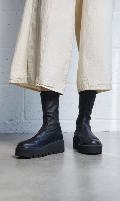 Plümo- Person wearing black Short Prato Boots and a cream skirt, standing on a concrete floor against a white wall.