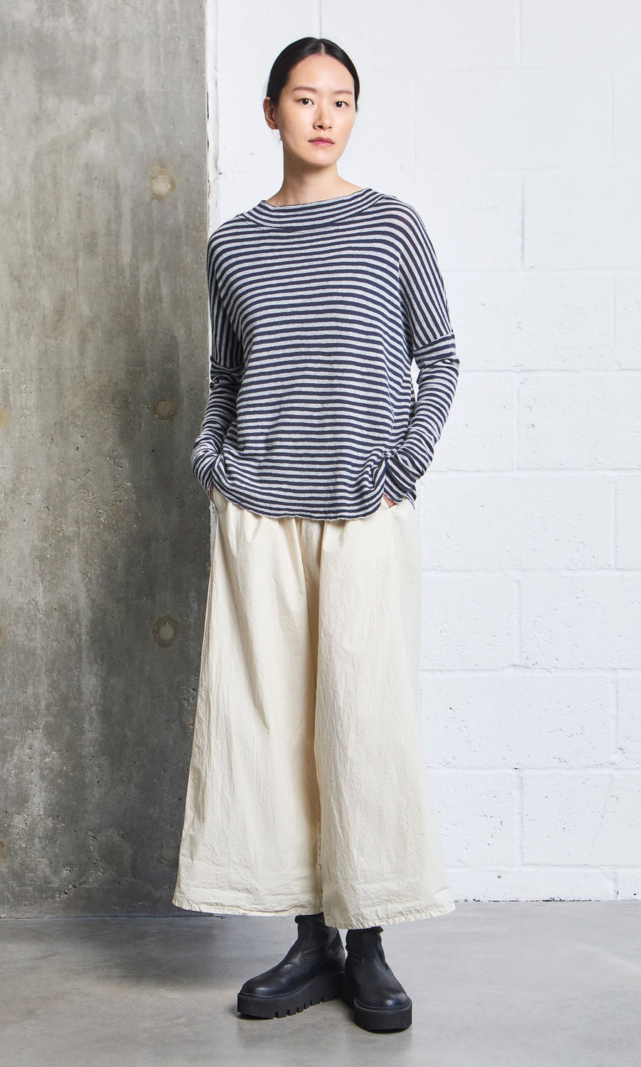 Plümo- Person wearing the Silvi Mockneck (dark navy stripe), wide cream pants, and black boots against a concrete wall.