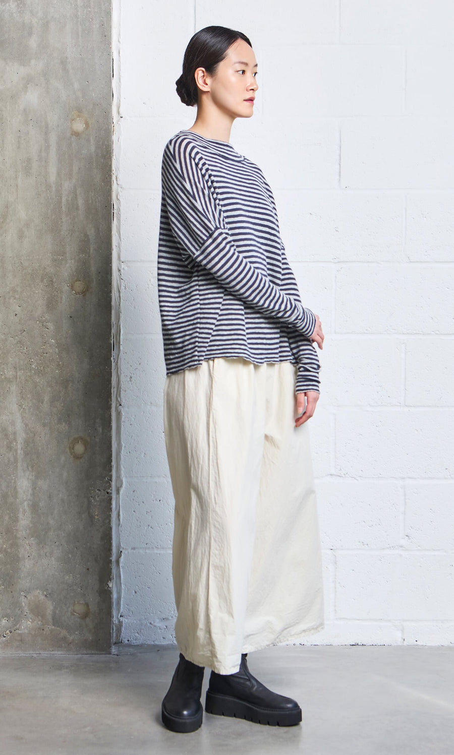 Plümo- A person stands sideways in the Silvi Mockneck (dark navy stripe), wide cream pants, and black boots by a concrete wall.