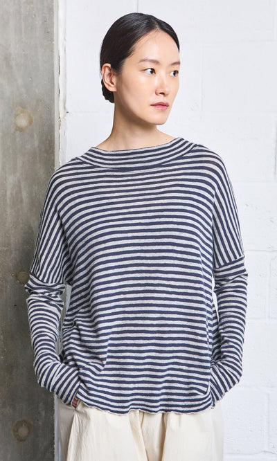 Plümo- Person with dark hair in a Silvi Mockneck (dark navy stripe) and light pants stands by a white and gray textured wall.