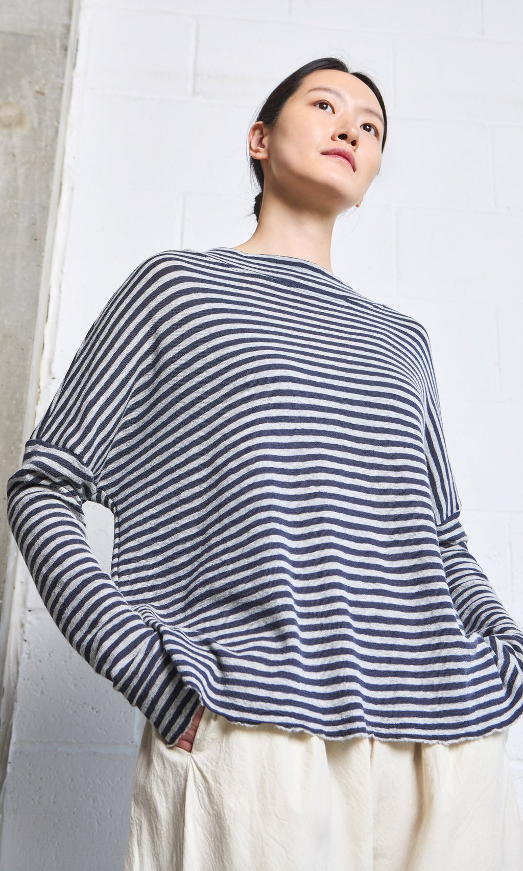 Plümo- Person wearing the Silvi Mockneck (dark navy stripe) and cream pants, standing against a white brick wall.