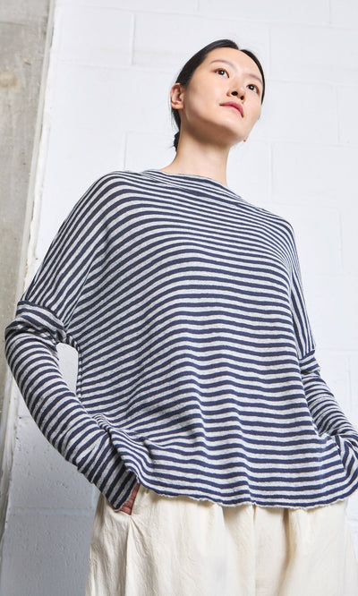 Plümo- Person wearing the Silvi Mockneck (dark navy stripe) and cream pants, standing against a white brick wall.