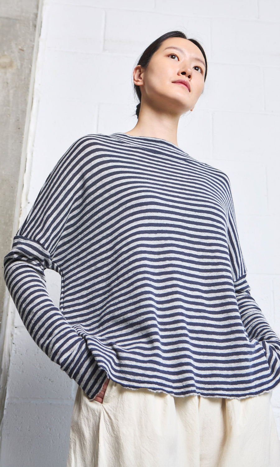 Plümo- Person wearing the Silvi Mockneck (dark navy stripe) and cream pants, standing against a white brick wall.