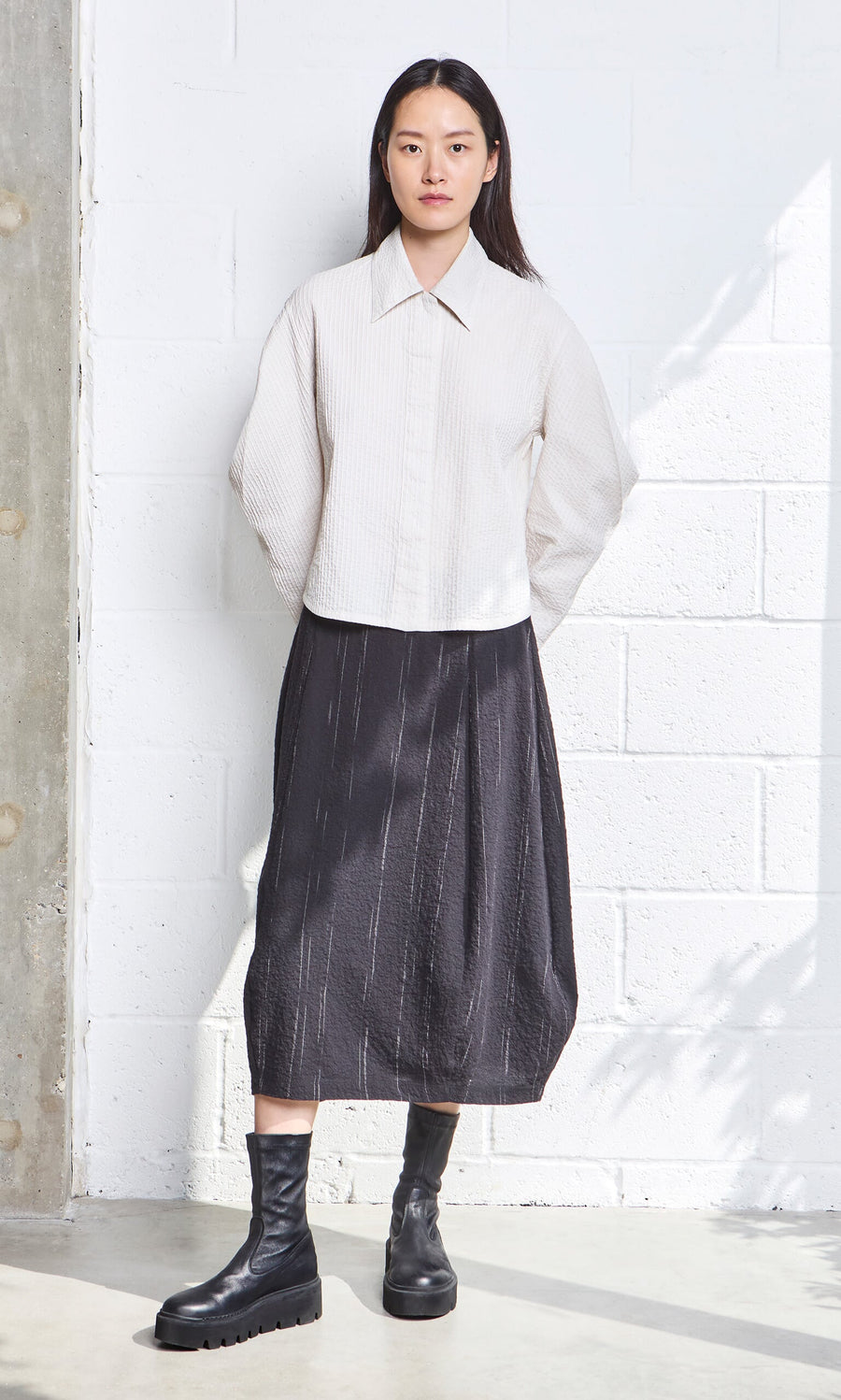 Plümo- Woman in a Ting Blouse with bishop sleeves, black midi skirt, and boots stands by a white brick wall.