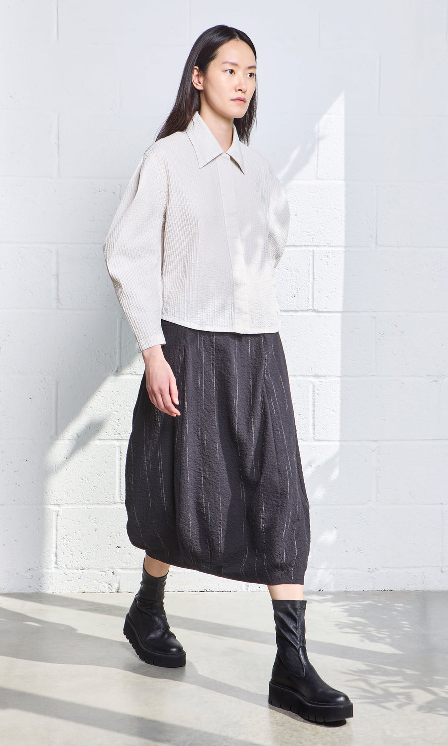 Plümo- Woman in a textured blouse, Speckle Skirt with elastic waist, and black boots by a sunlit white brick wall.