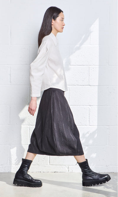 Plümo- A woman in a white blouse and the Speckle Skirt walks past a white textured wall in bright natural light.
