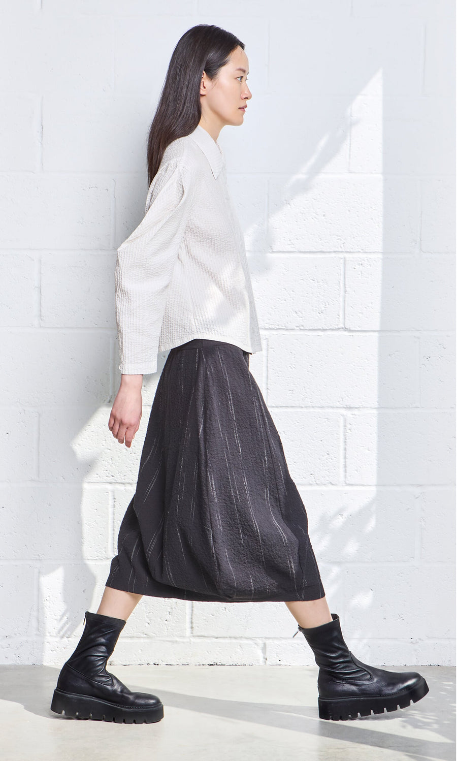 Plümo- A woman in a white blouse and the Speckle Skirt walks past a white textured wall in bright natural light.