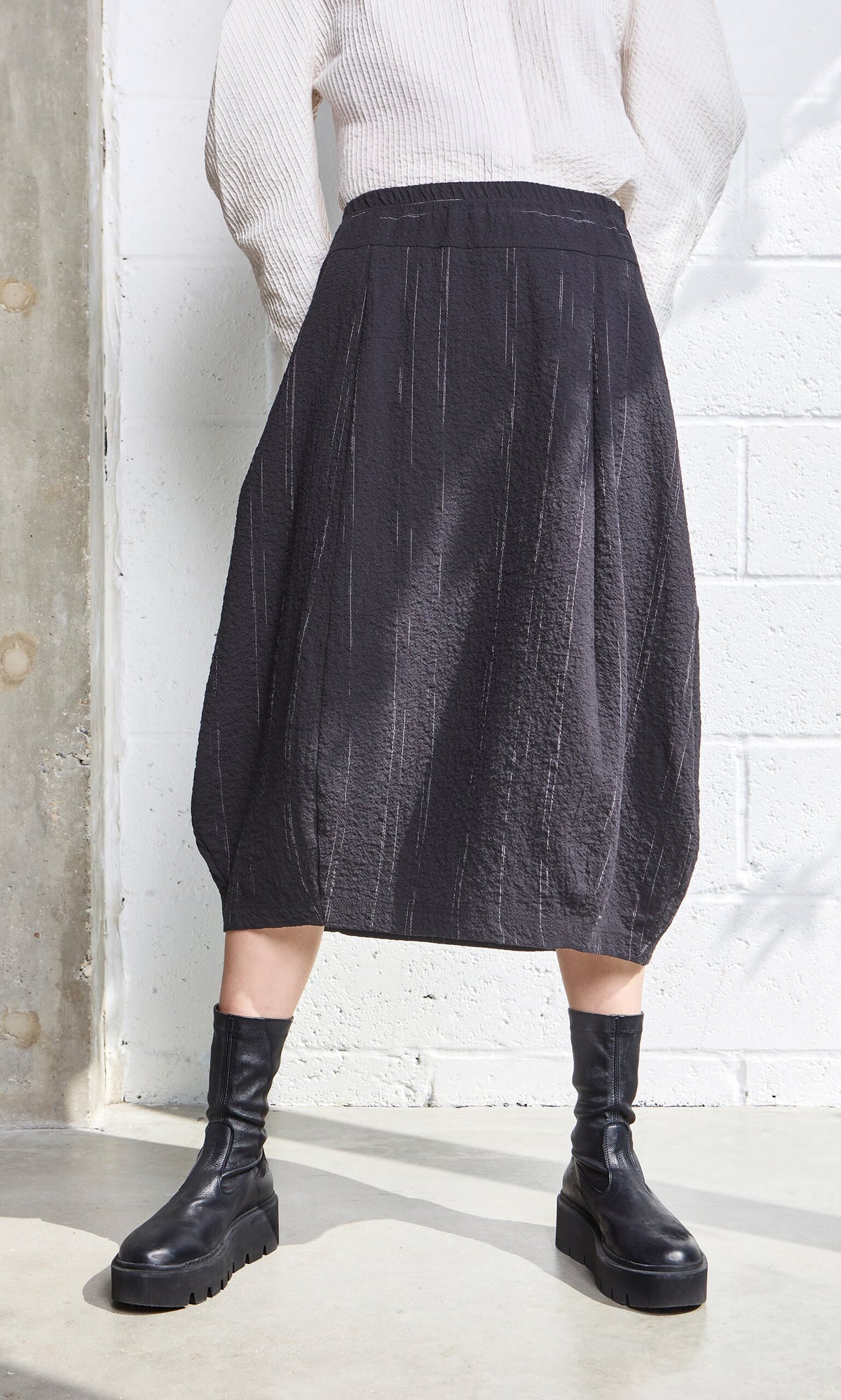 Plümo- Person in a Speckle Skirt with elastic waist, black boots, and white top stands by a white wall with concrete details.