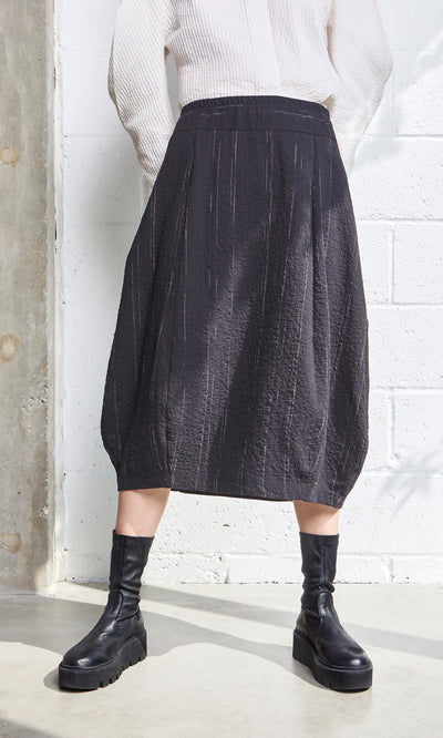 Plümo- Person in a Speckle Skirt with elastic waist, black boots, and white top stands by a white wall with concrete details.