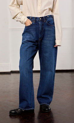 Plümo- Person wearing Stack Jeans, cream button-up shirt, and black shoes standing indoors on a dark floor.