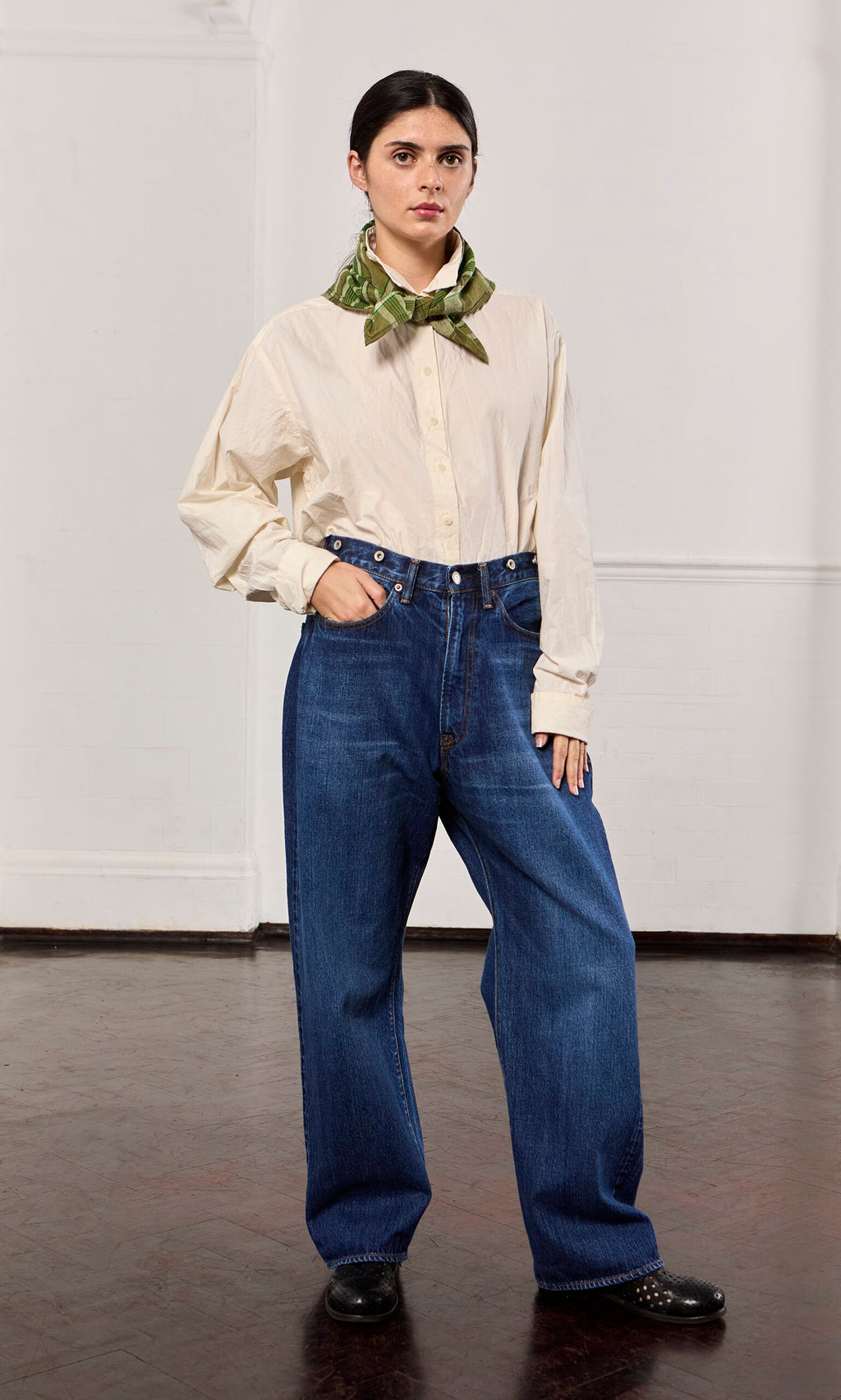 Plümo- Woman in loose cream shirt, green scarf, and Stack Jeans stands in a plain indoor setting.