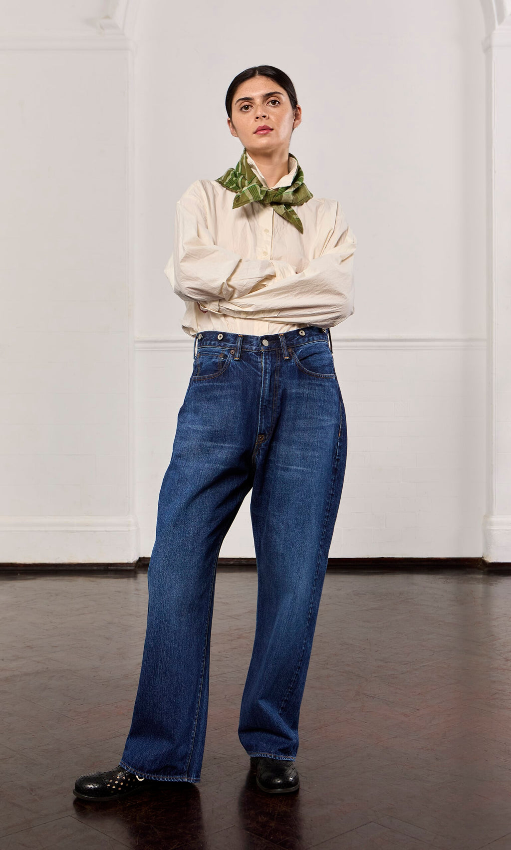 Plümo- Woman with arms crossed wears beige shirt, green scarf, black shoes, and Stack Jeans in a plain room.