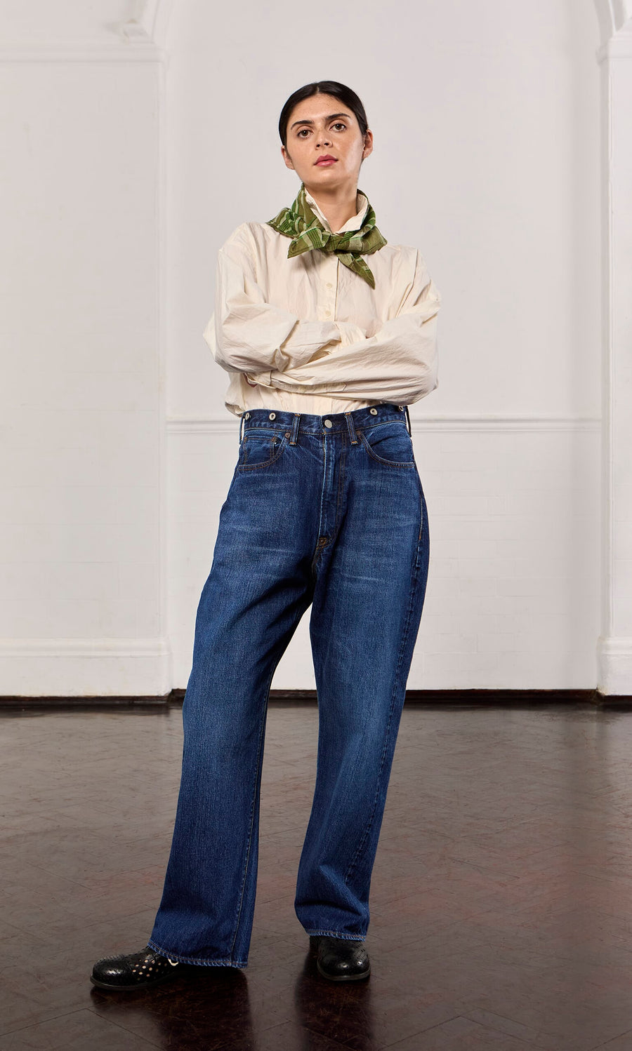 Plümo- Woman with arms crossed wears beige shirt, green scarf, black shoes, and Stack Jeans in a plain room.