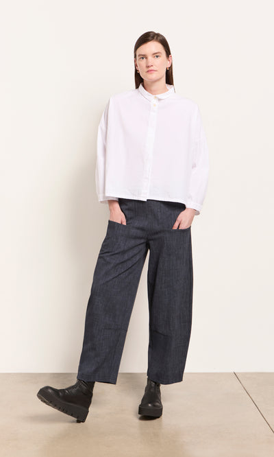 Plümo  Woman in a white button-up, Stanner Pants featuring a curved leg silhouette, and black boots against a plain background.