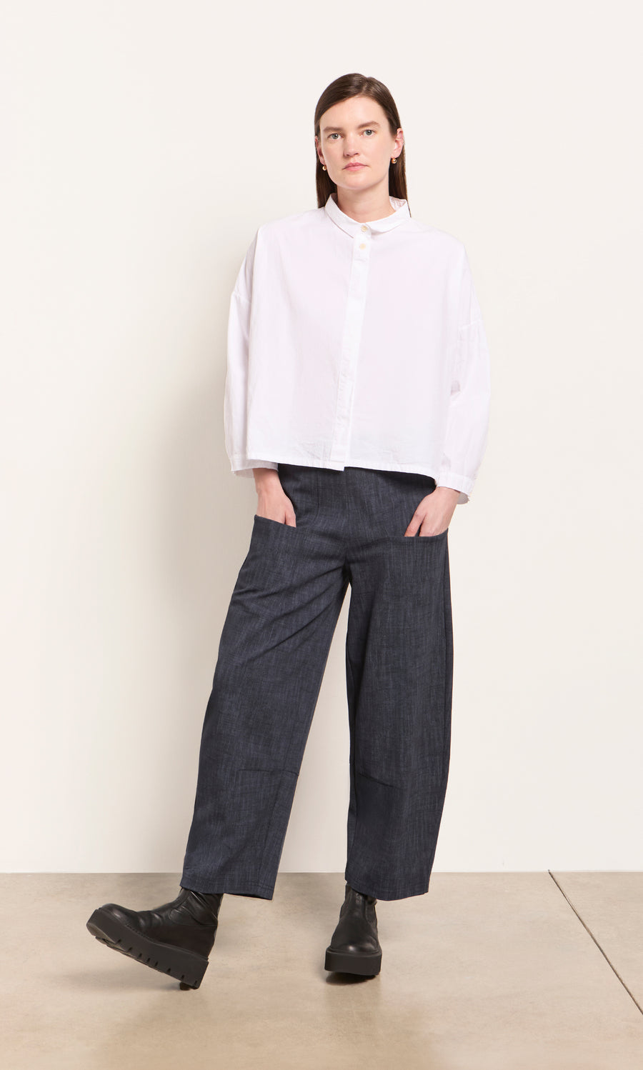 Plümo  Woman in a white button-up, Stanner Pants featuring a curved leg silhouette, and black boots against a plain background.