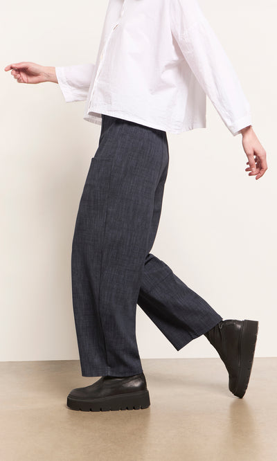 Plümo  Person wearing Stanner Pants with a curved leg, a white shirt, and black platforms walks on a tan floor.