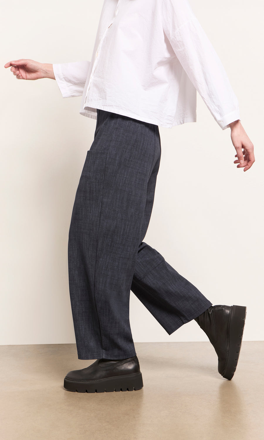 Plümo  Person wearing Stanner Pants with a curved leg, a white shirt, and black platforms walks on a tan floor.