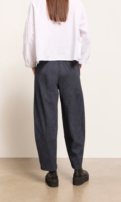 Plümo  Person wearing Stanner Pants—indigo denim with a curved leg, comfortable fit, black shoes, on a beige floor.