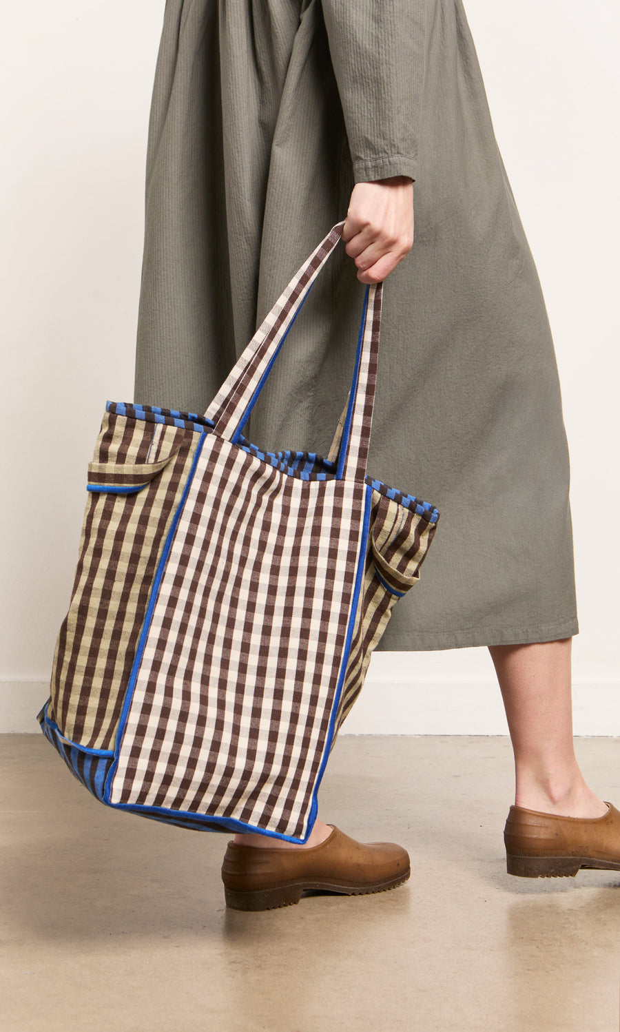 Plümo- Person in a long gray dress and brown shoes holding the Stylist Bag, a spacious blue and brown gingham tote.