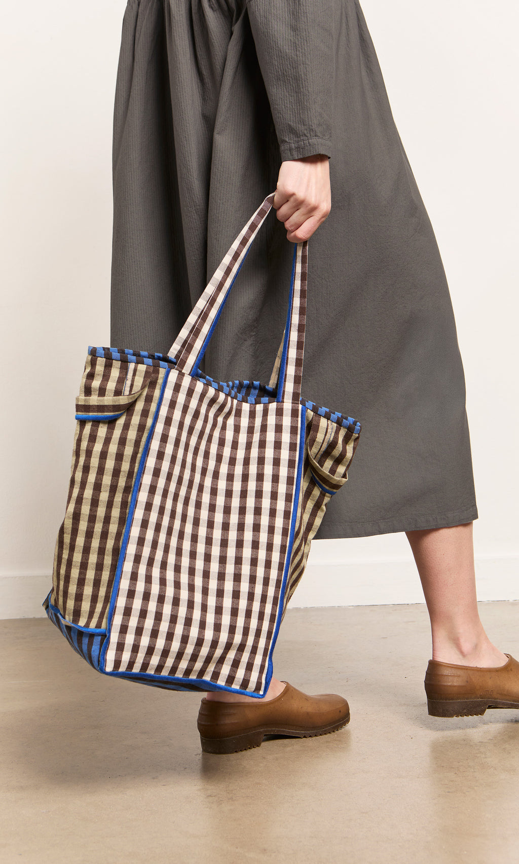Plümo  Person in a gray dress holds Stylist Bag with shoulder straps, wearing brown clogs on a light floor.