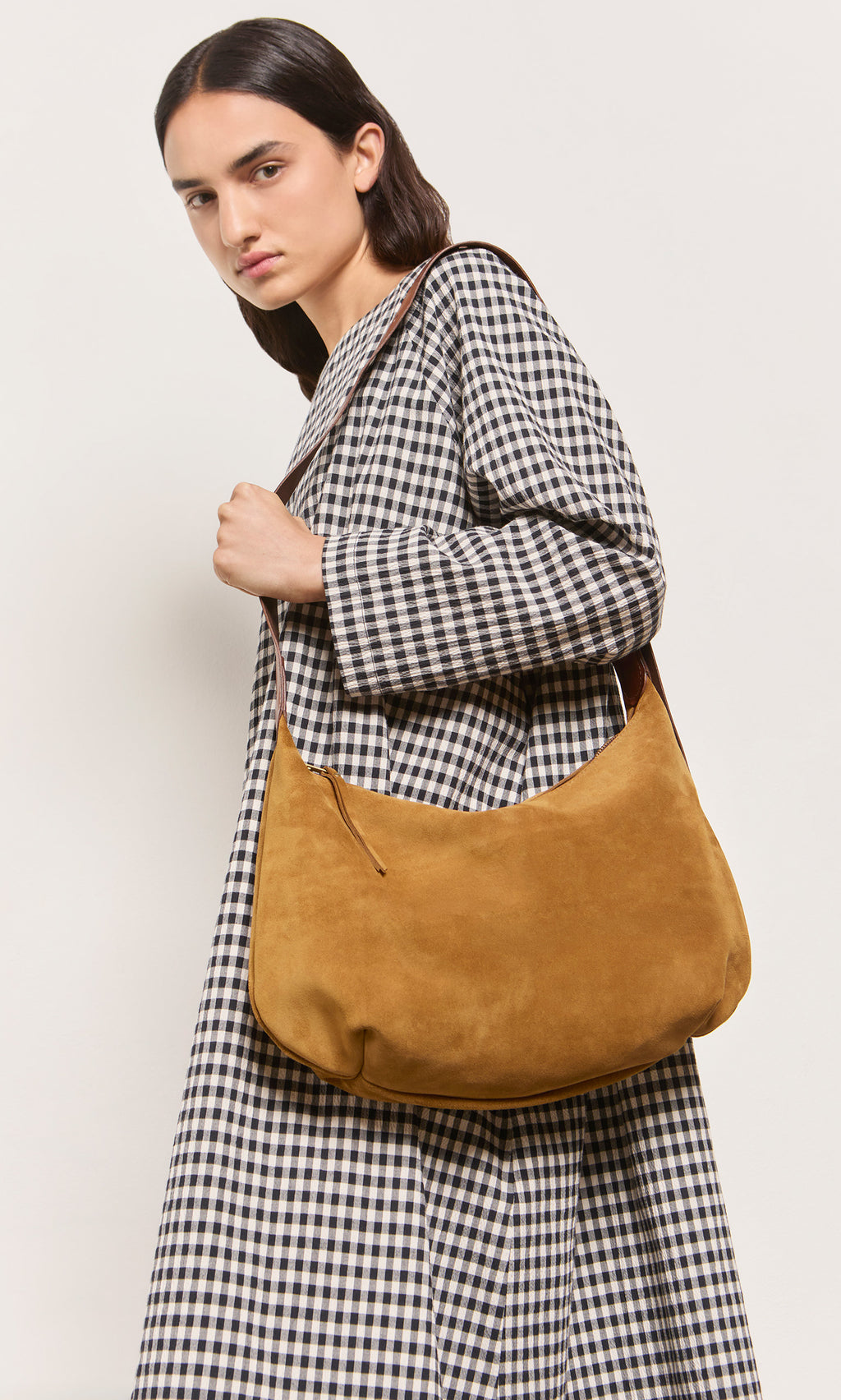 Plümo  Person in a gingham dress holds the Kiel Suede Bag, gazing confidently at the camera.