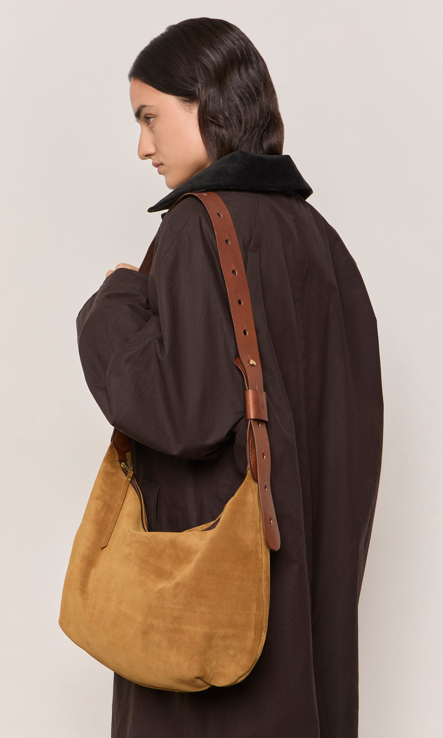 Plümo  Person in a dark coat with a tan Kiel Suede Bag with brown leather strap, facing left.