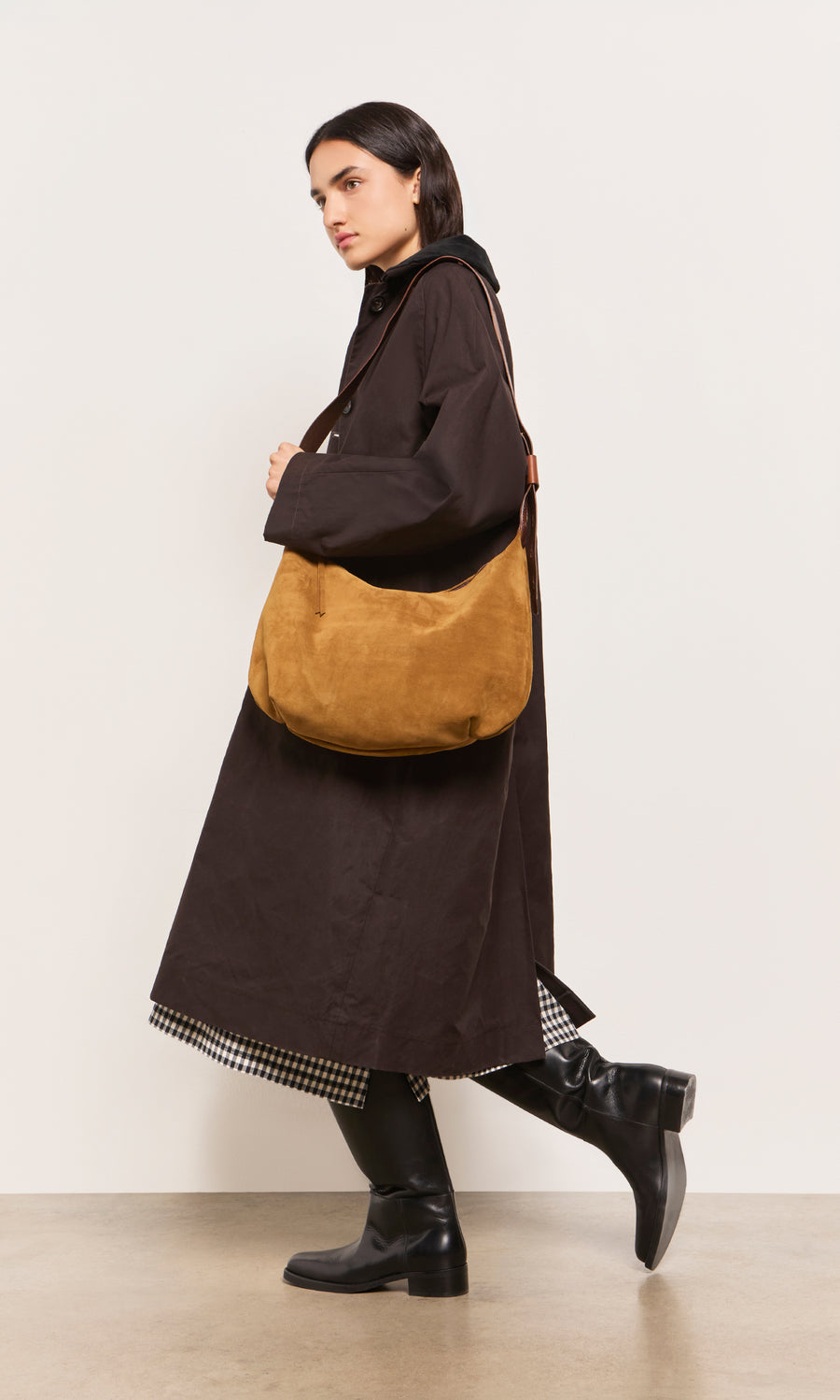 Plümo  Woman in a dark trench coat, black boots, and Kiel Suede Bag walking against a plain backdrop.