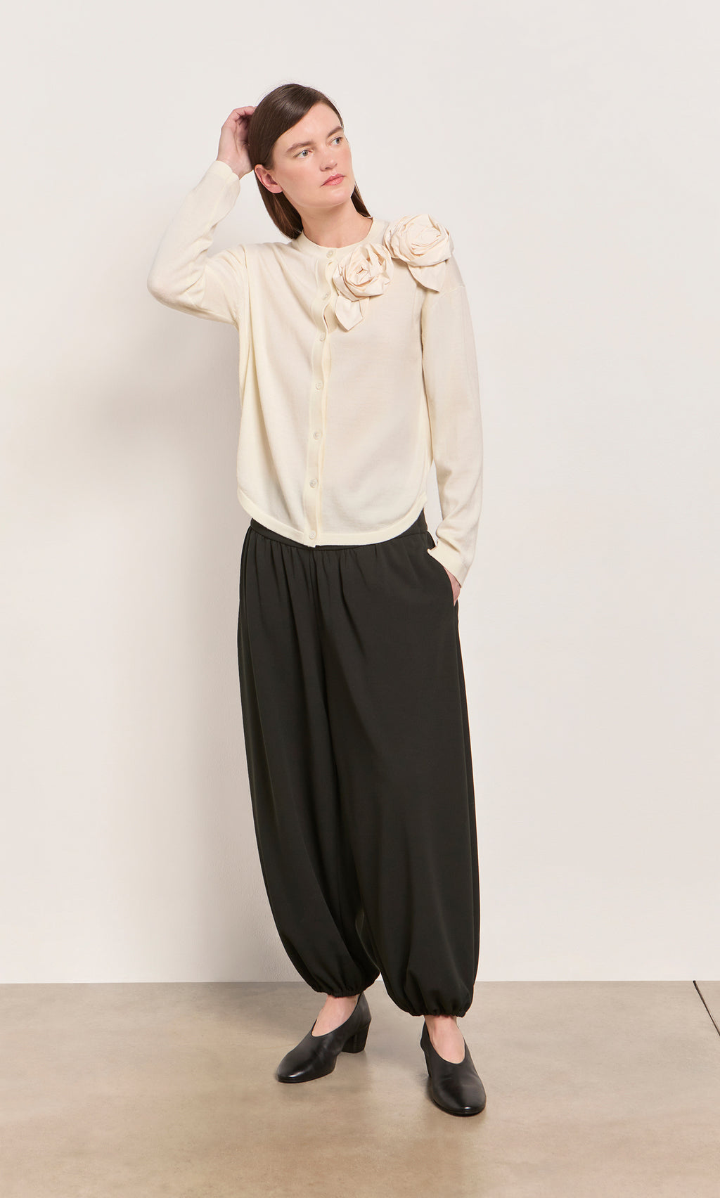 Plümo  Woman in the Sugared Corsage Cardigan and loose black pants stands on a light floor, looking to the side.
