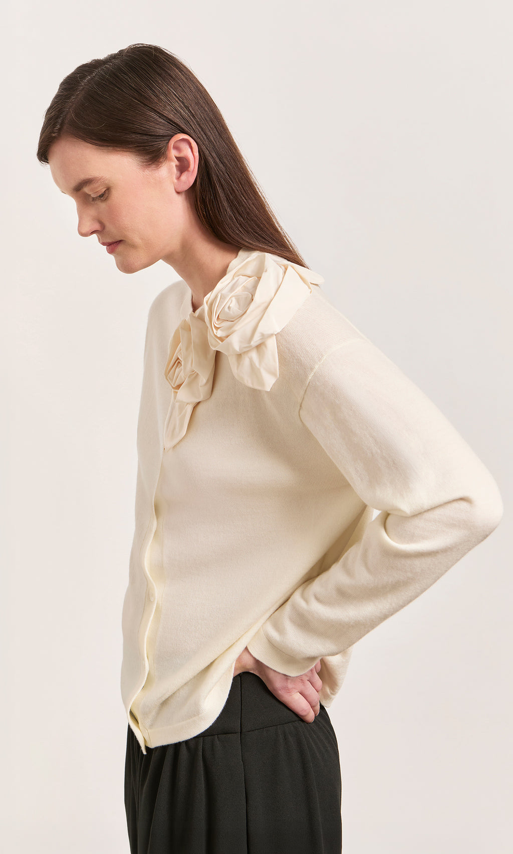 Plümo  Woman in Sugared Corsage Cardigan with sculptural shoulder corsages, standing sideways, hands on hips.