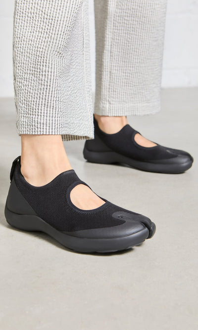 Plümo- Person wears Tabi Sandals in black with white textured pants, standing on a light-colored floor.
