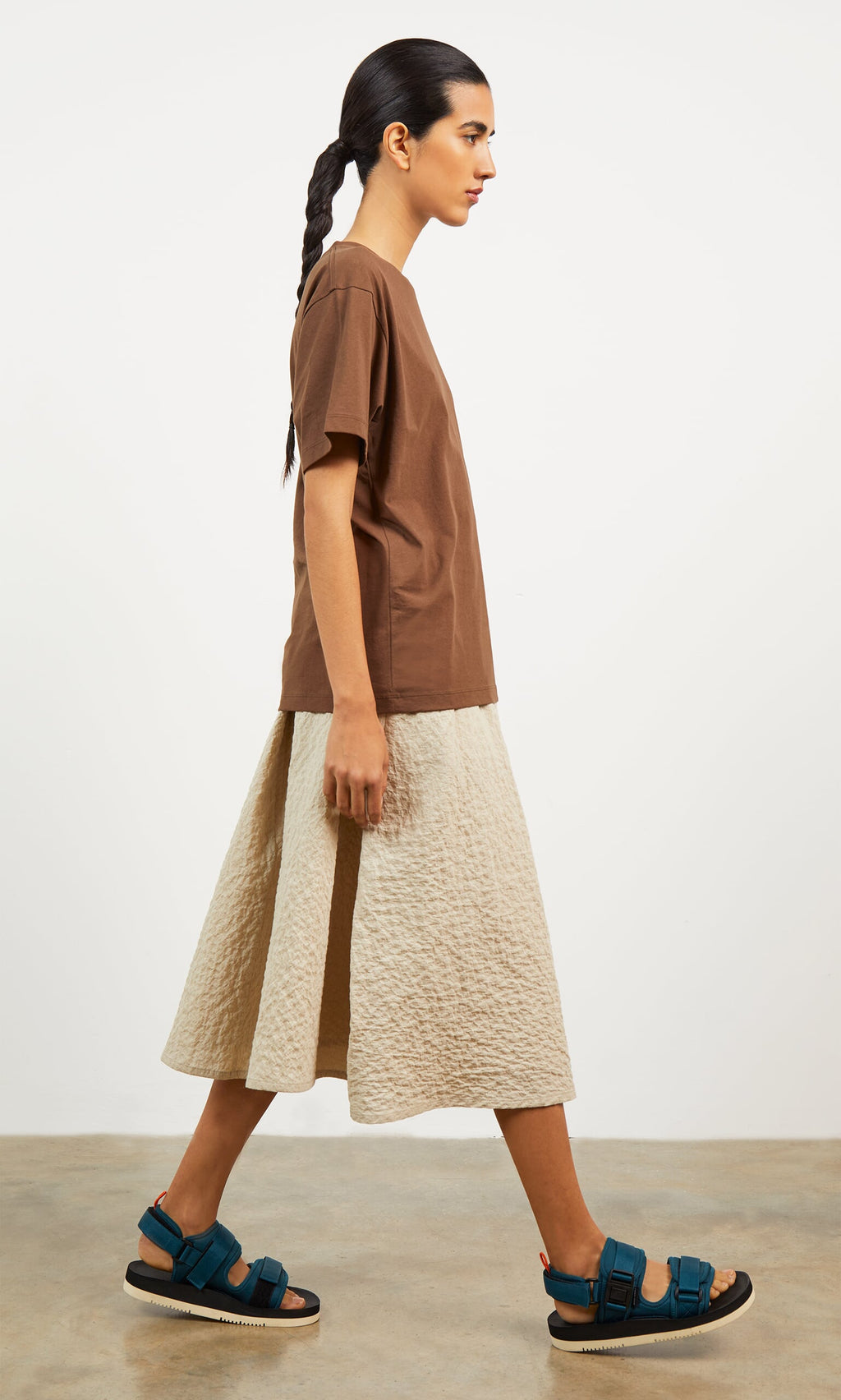 Plümo- Woman in a brown t-shirt, Jane Skirt with asymmetric pleating, and blue sandals walks on a concrete floor.