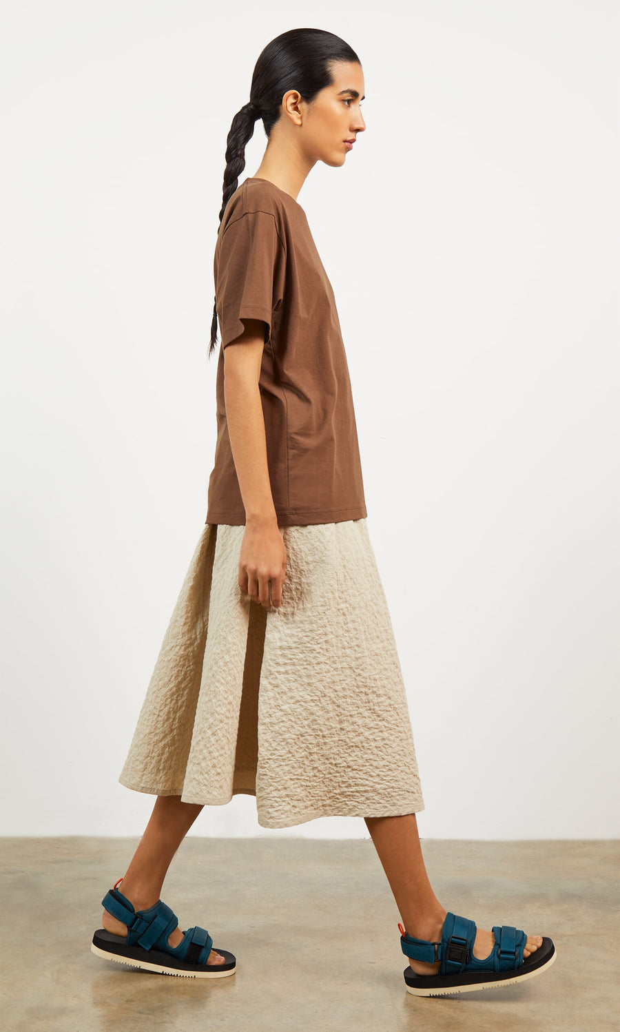 Plümo- Woman in a brown t-shirt, Jane Skirt with asymmetric pleating, and blue sandals walks on a concrete floor.