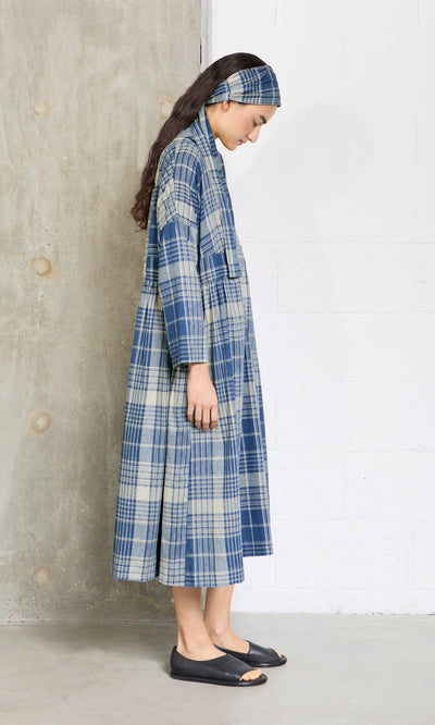 Plümo- Woman in a Tarkhan Dress - Check and matching headband stands sideways against a concrete and white wall.