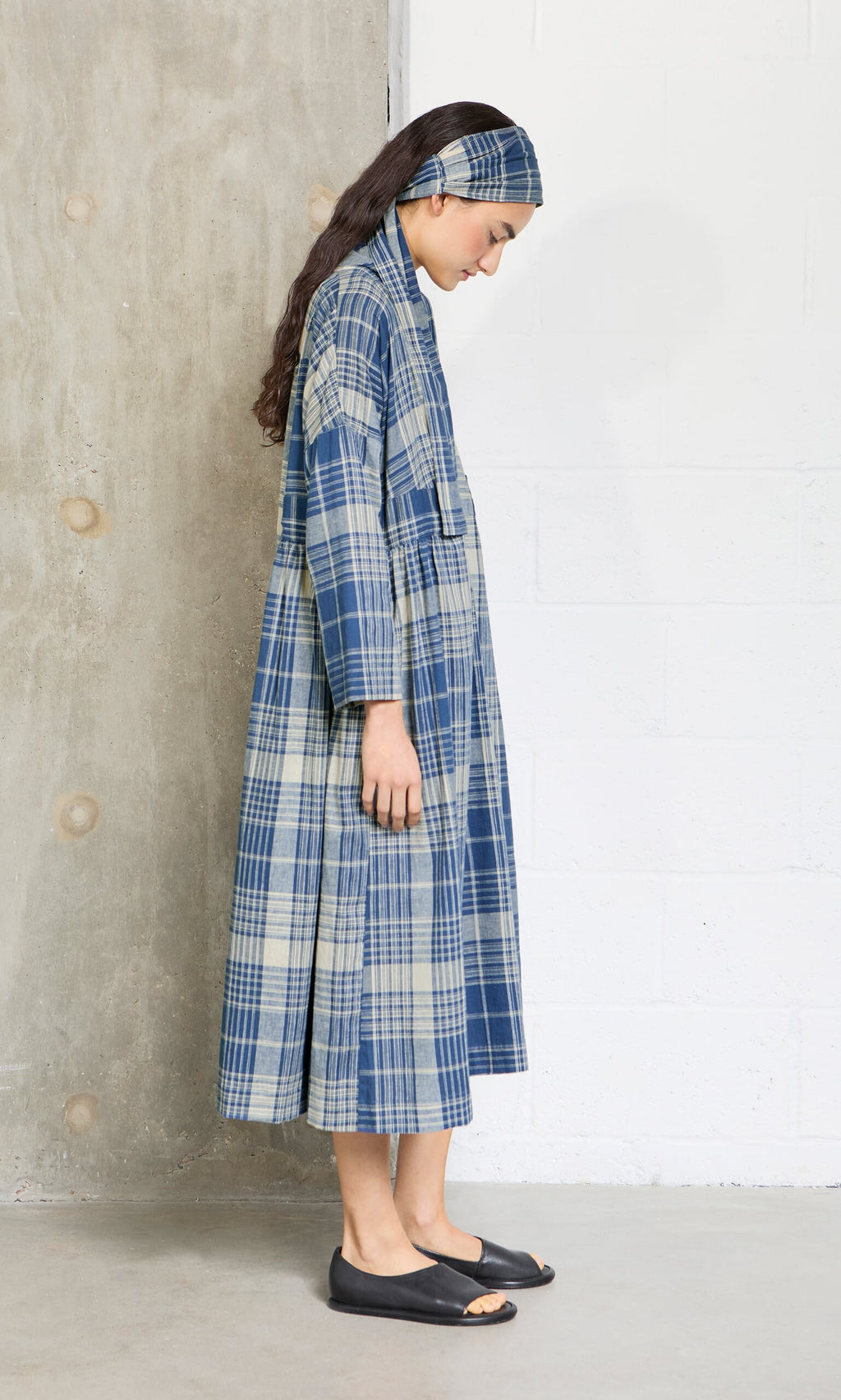Plümo- Woman in the Wrap Tie - Check, a long blue plaid dress with extra-long belt, standing sideways in black flats.