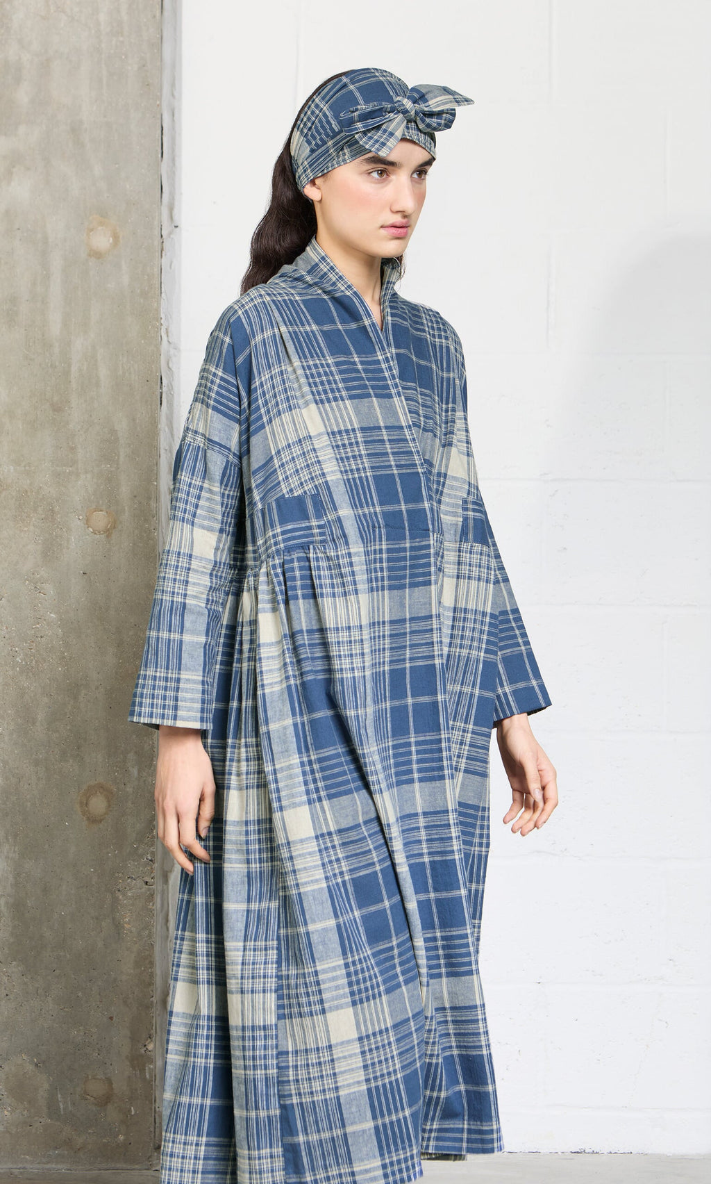 Plümo- Woman in the Wrap Tie - Check dress and matching headscarf, made from Japanese Banshu-ori fabric by a concrete wall.