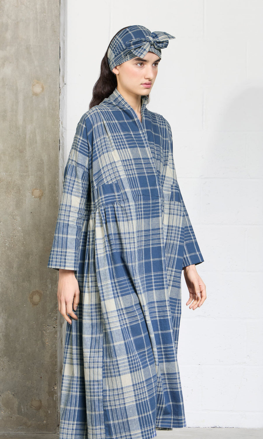 Plümo- Woman in the Wrap Tie - Check dress and matching headscarf, made from Japanese Banshu-ori fabric by a concrete wall.