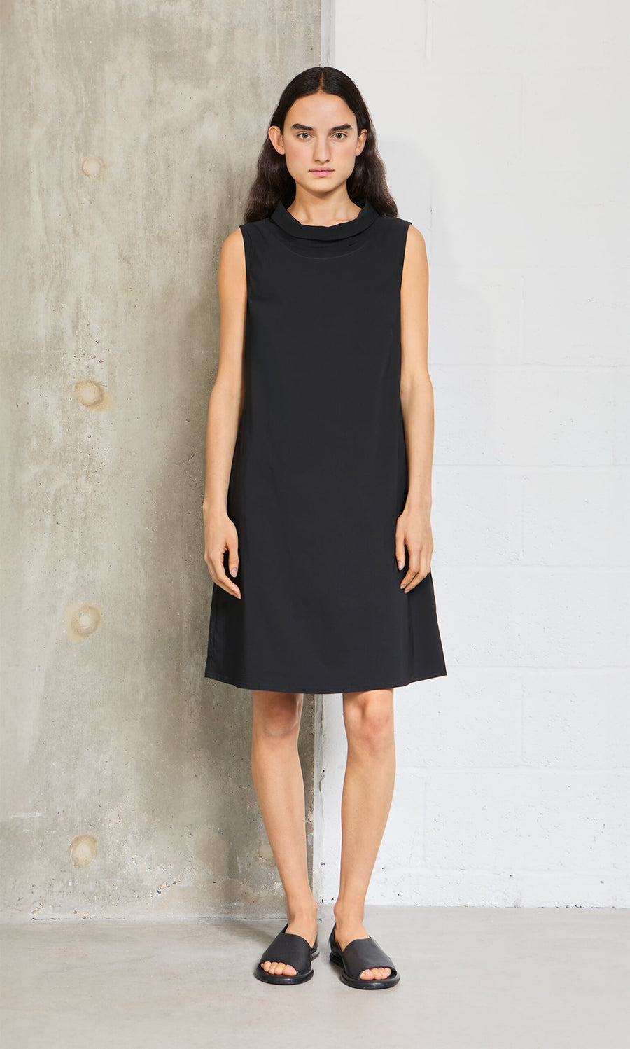 Plümo- A person stands indoors in the Tegan Dress and sandals, set against a concrete and white brick wall.