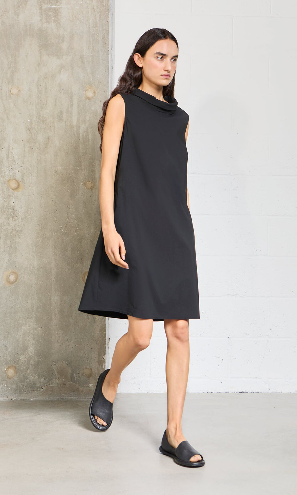 Plümo- Person wearing the Tegan Dress and black sandals, walking indoors near a concrete and white brick wall.
