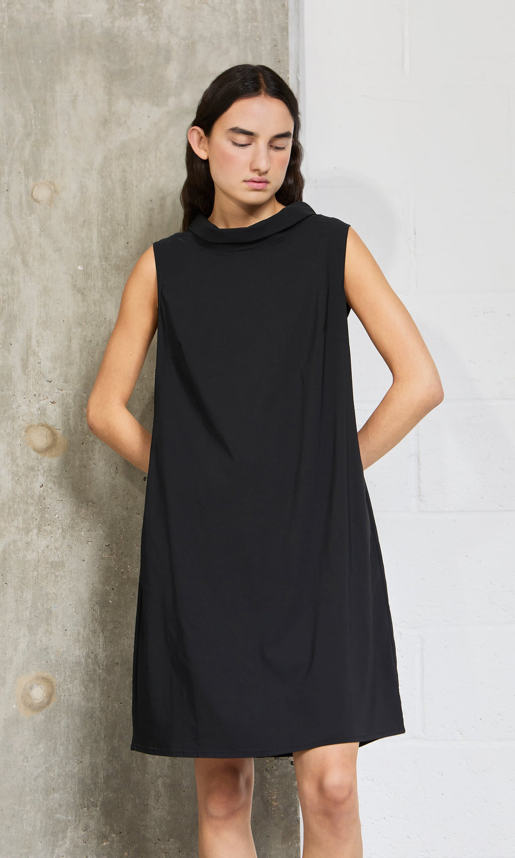 Plümo- Someone wearing the Tegan Dress stands against a concrete wall, looking down with hands behind their back.