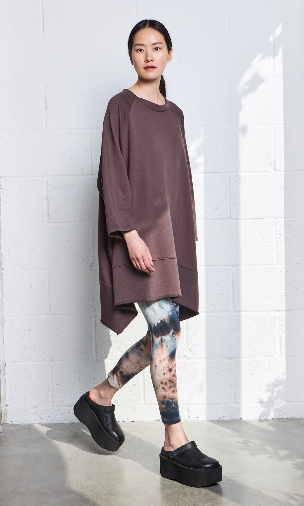 Plümo- Woman in a relaxed fit Kelie Sweater, tie-dye leggings, and black platform shoes stands by a white brick wall.