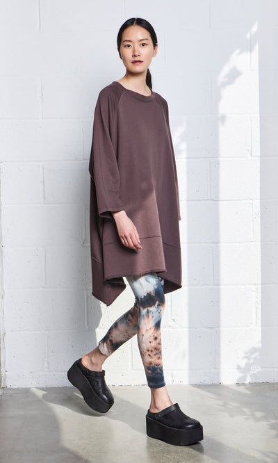 Plümo- A woman stands by a white brick wall in a loose brown tunic, tie-dye leggings, and black Kim Clogs in sunlight.